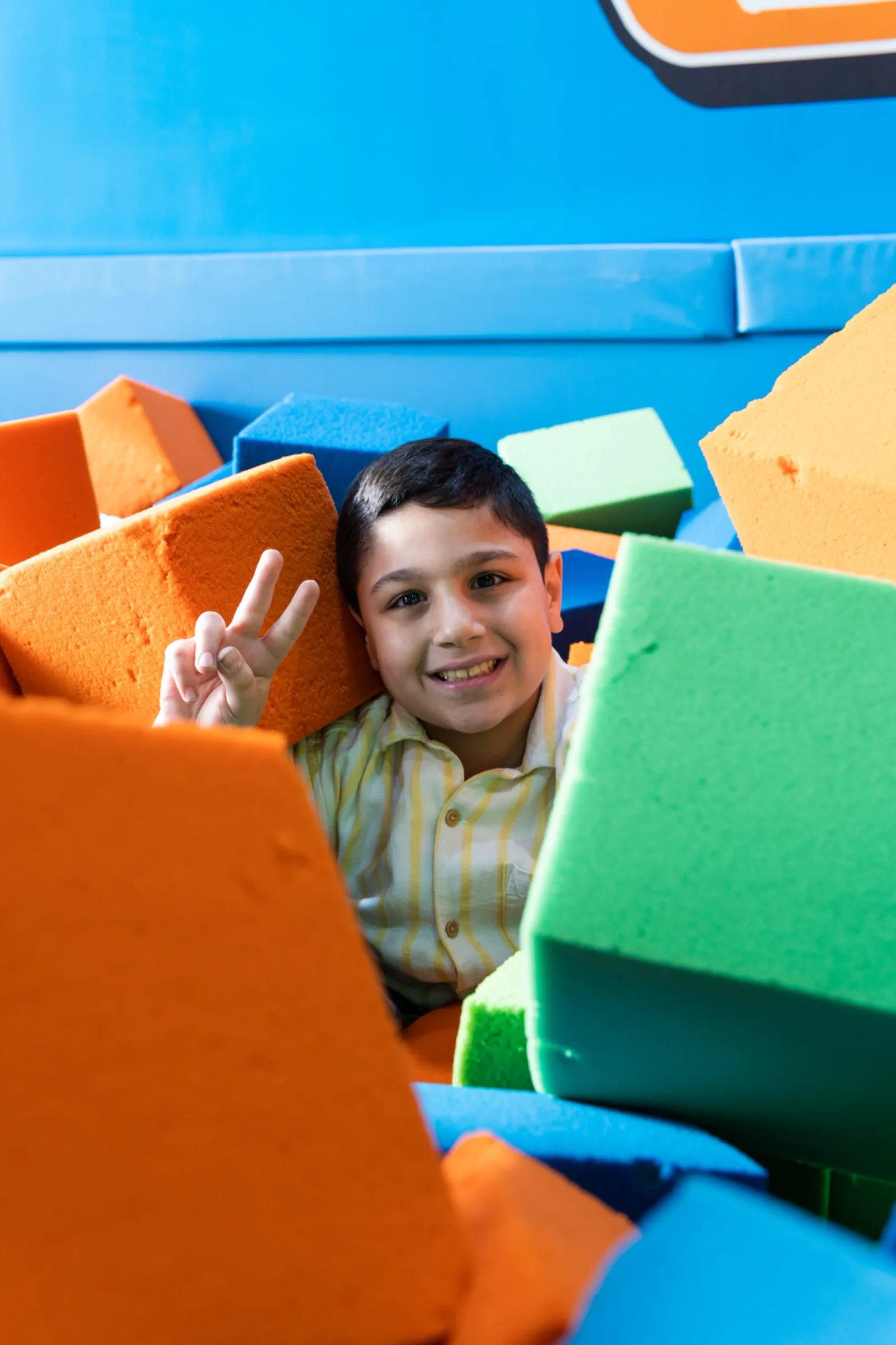 Smiling boy buried in colorful foam cubes flashing a peace sign at Trampo Kuwait