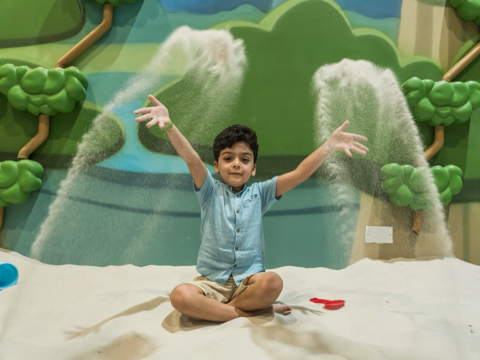 Boy laughing as he tosses two big handfuls of sand into the air, painted forest backdrop behind him