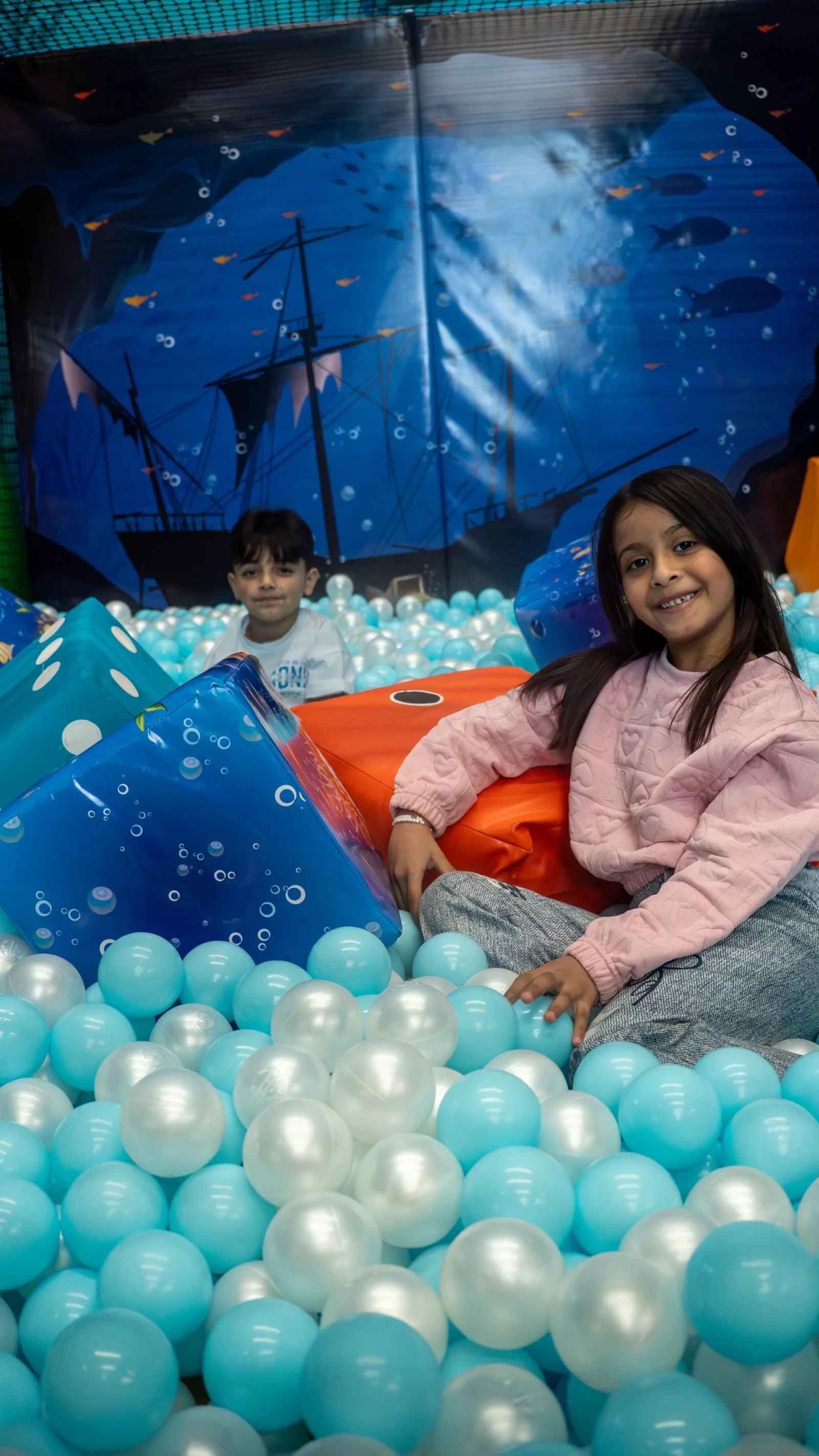 Brother and sister relaxing among pearl-blue balls in front of the pirate-ship soft play backdrop