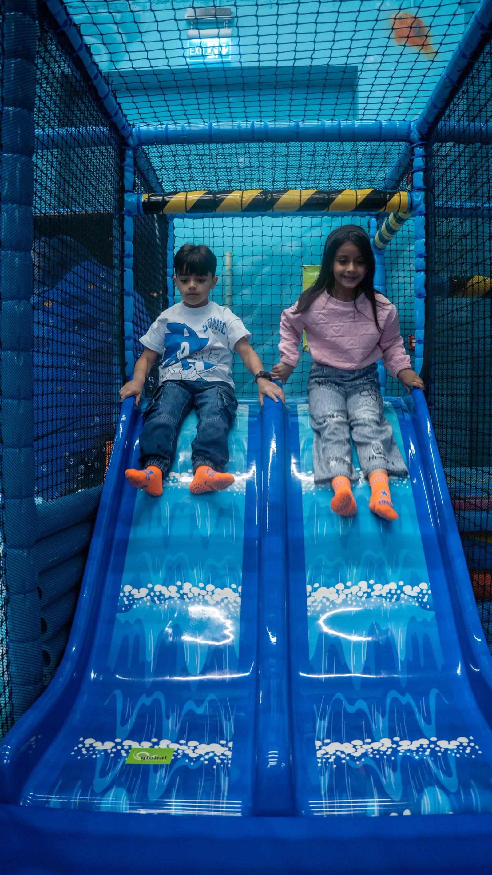 Two kids in orange grip socks gliding side by side down the gentle twin slide