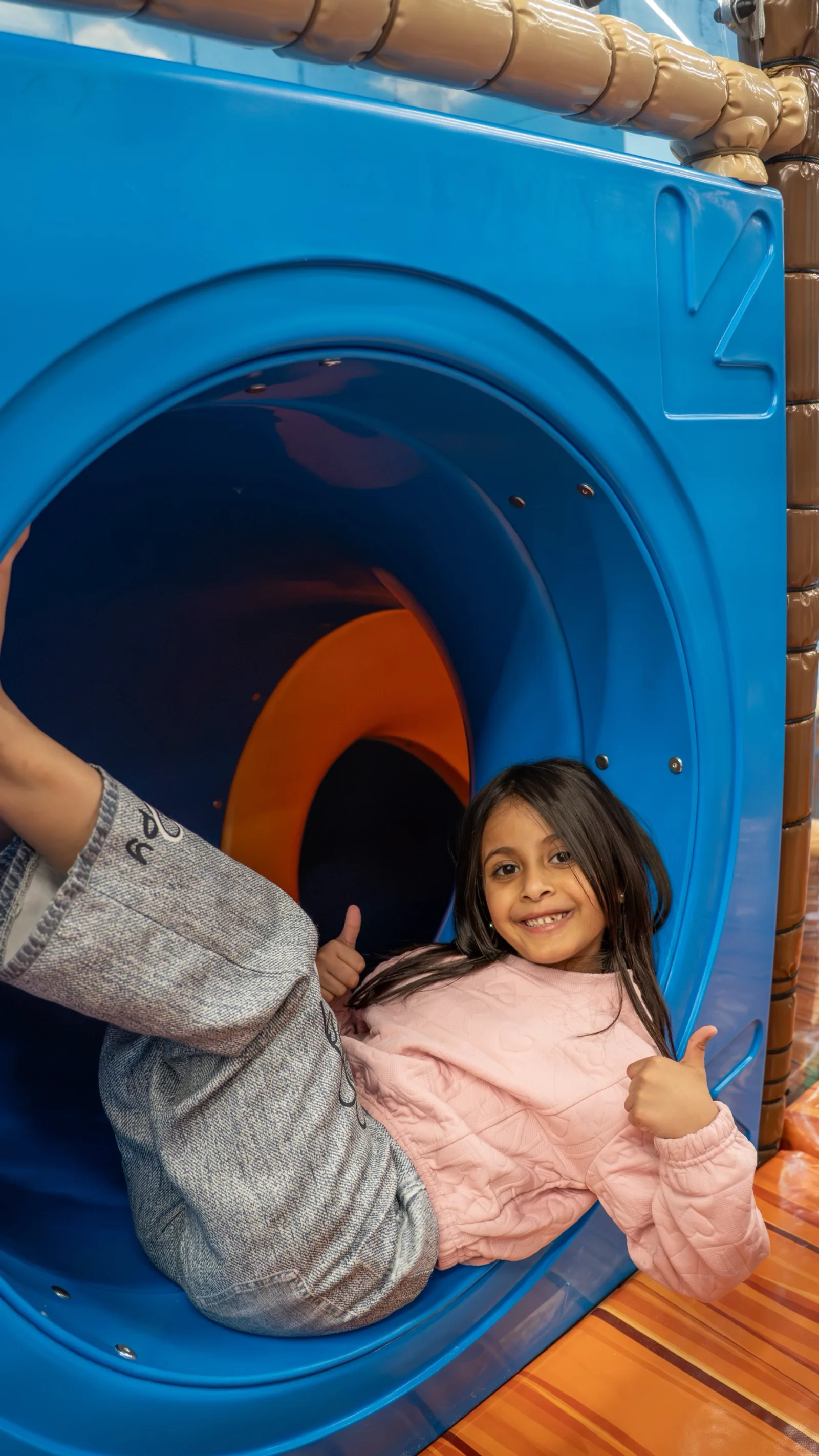 Smiling girl giving a thumbs up at the entrance of the round blue tunnel slide