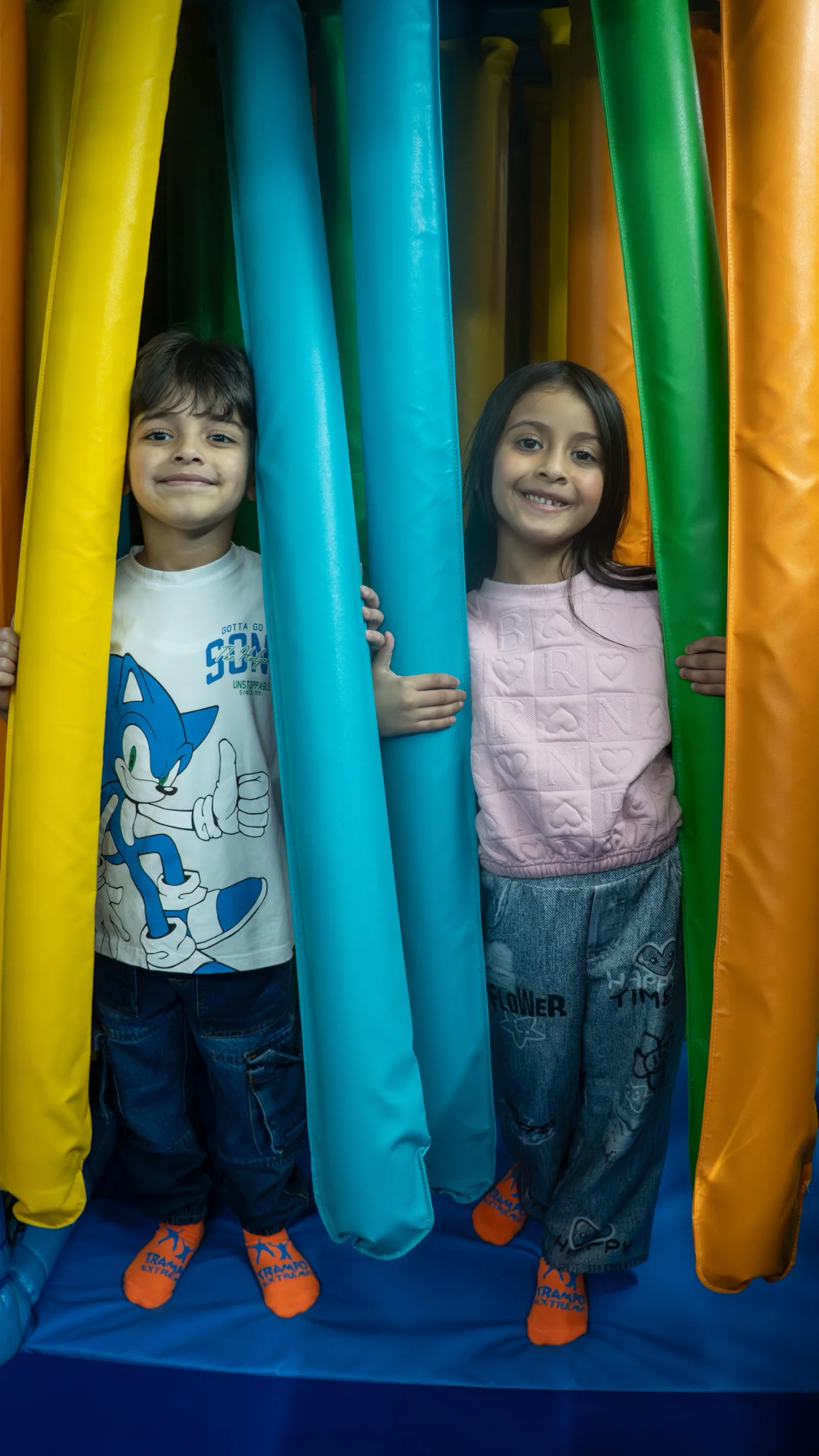 Boy and girl peeking through the colourful padded curtain columns of the soft play tunnel