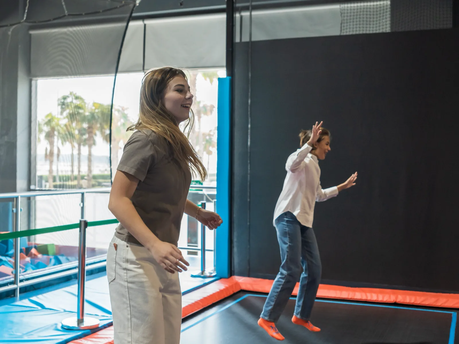 Two friends bouncing in sync on the Valo Jump trampolines, sea-front palm trees through the window